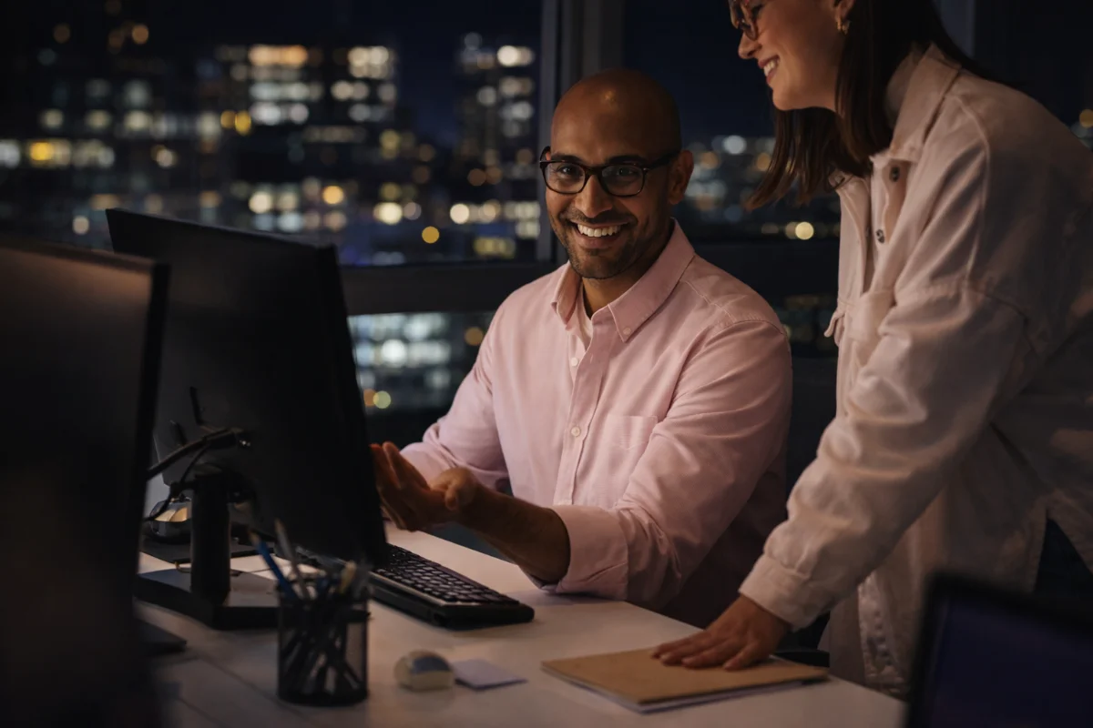 Colegas de trabalho sorrindo em frente ao computador à noite, fundo iluminado da cidade.