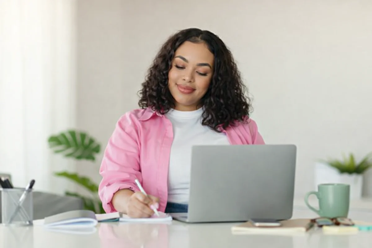 Mulher jovem sorrindo, olhos fechados, escrevendo em um caderno com laptop na mesa.