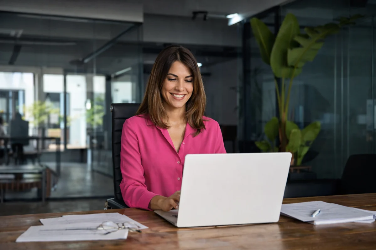 Mulher sorridente em camisa rosa no escritório, digitando em notebook branco.