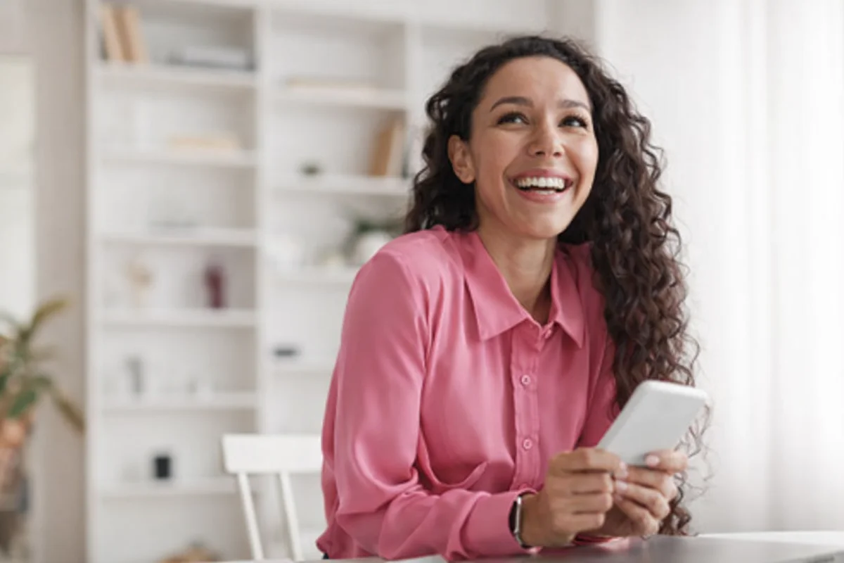 Mulher sorridente de cabelo cacheado em camisa rosa, olhando para cima e segurando um celular.