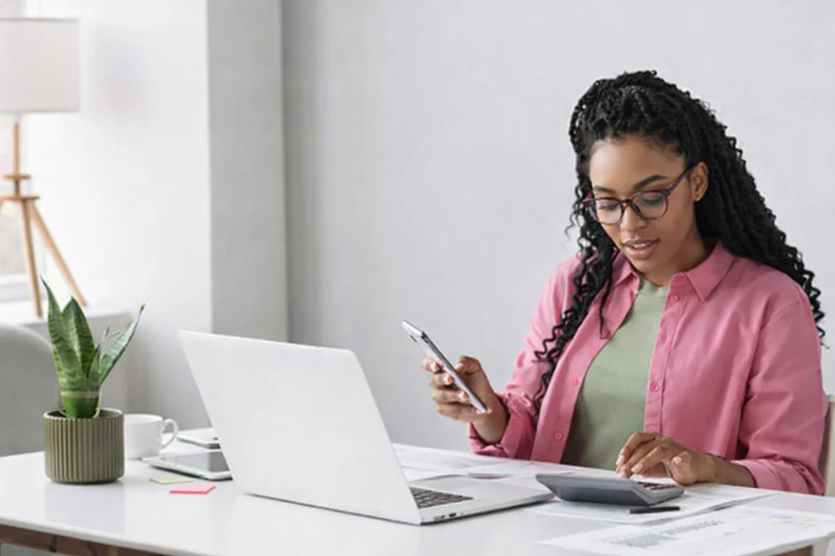 Mulher trabalha em casa com laptop e calculadora, organizando finanças.