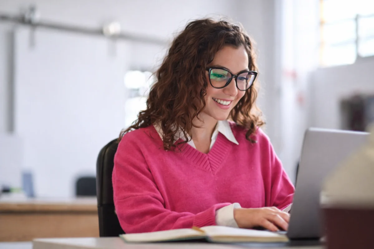 Mulher jovem sorri usando óculos e suéter rosa, trabalhando em notebook.