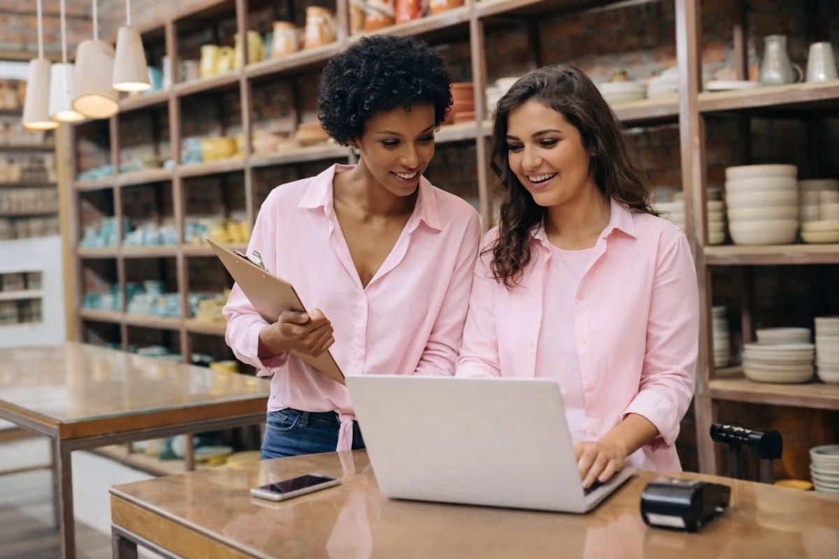 Duas mulheres sorrindo gerenciando estoques com laptop em loja de artesanato.