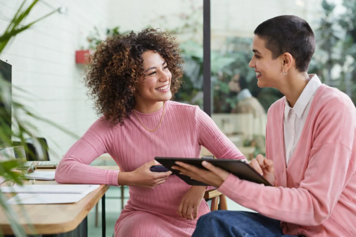 Duas mulheres sorrindo, discutindo tablet em escritório.