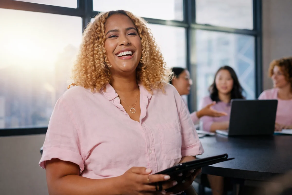 Mulher de cabelos cacheados sorrindo, segurando tablet, com colegas em escritório.