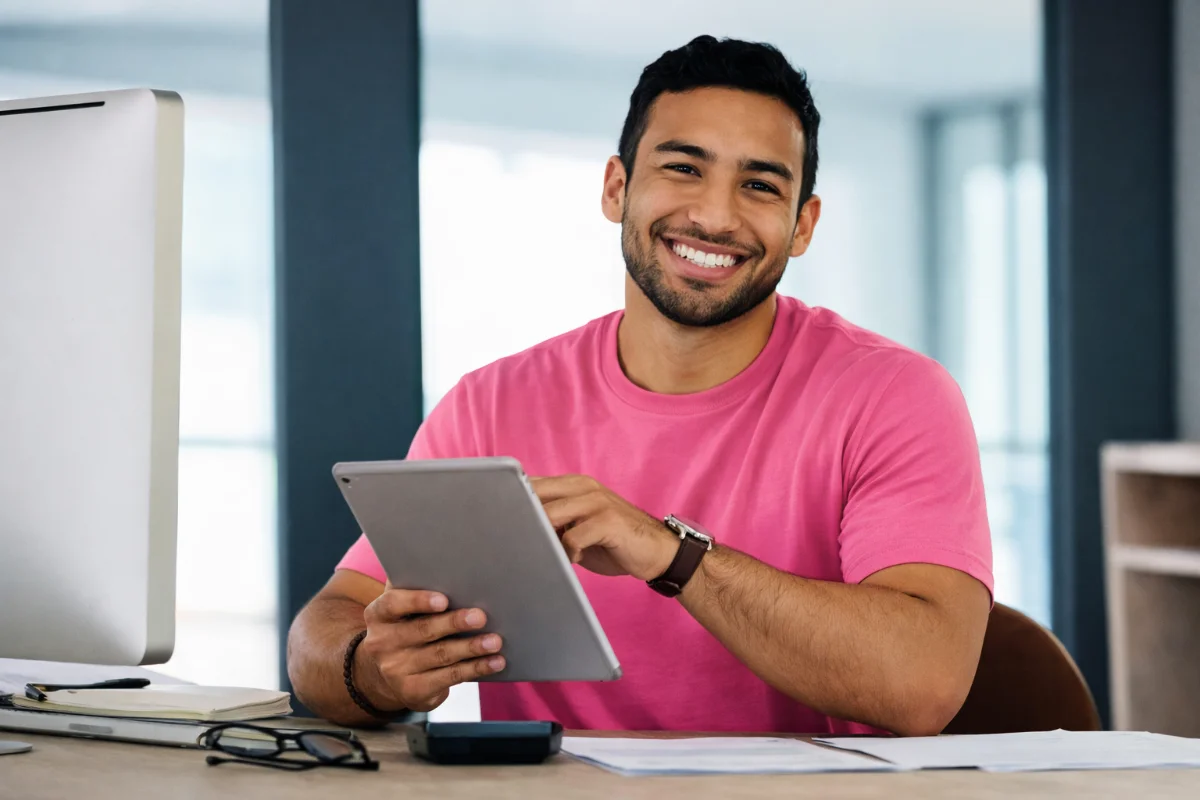 Homem feliz de camiseta rosa usando tablet e sorrindo em seu escritório, com monitor ao fundo.