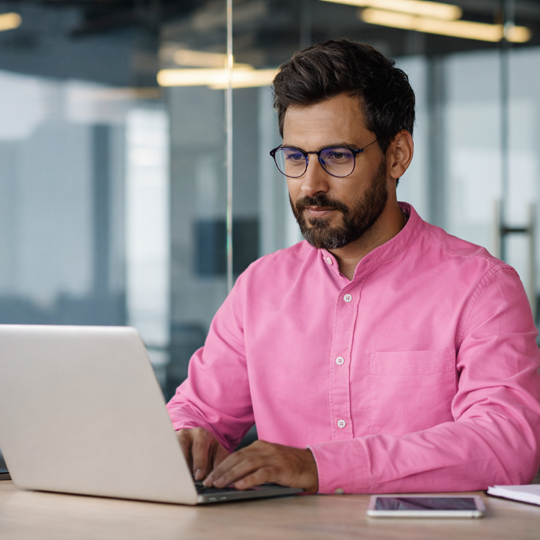 Homem de barba, óculos e camisa rosa trabalhando em notebook numa mesa.