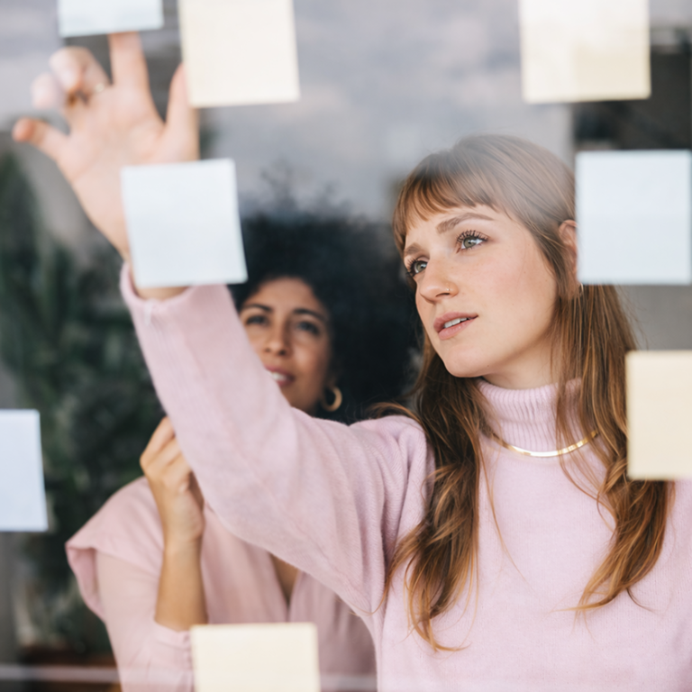 Mulheres estrategistas colando post-its em vidro, brainstorming colaborativo.